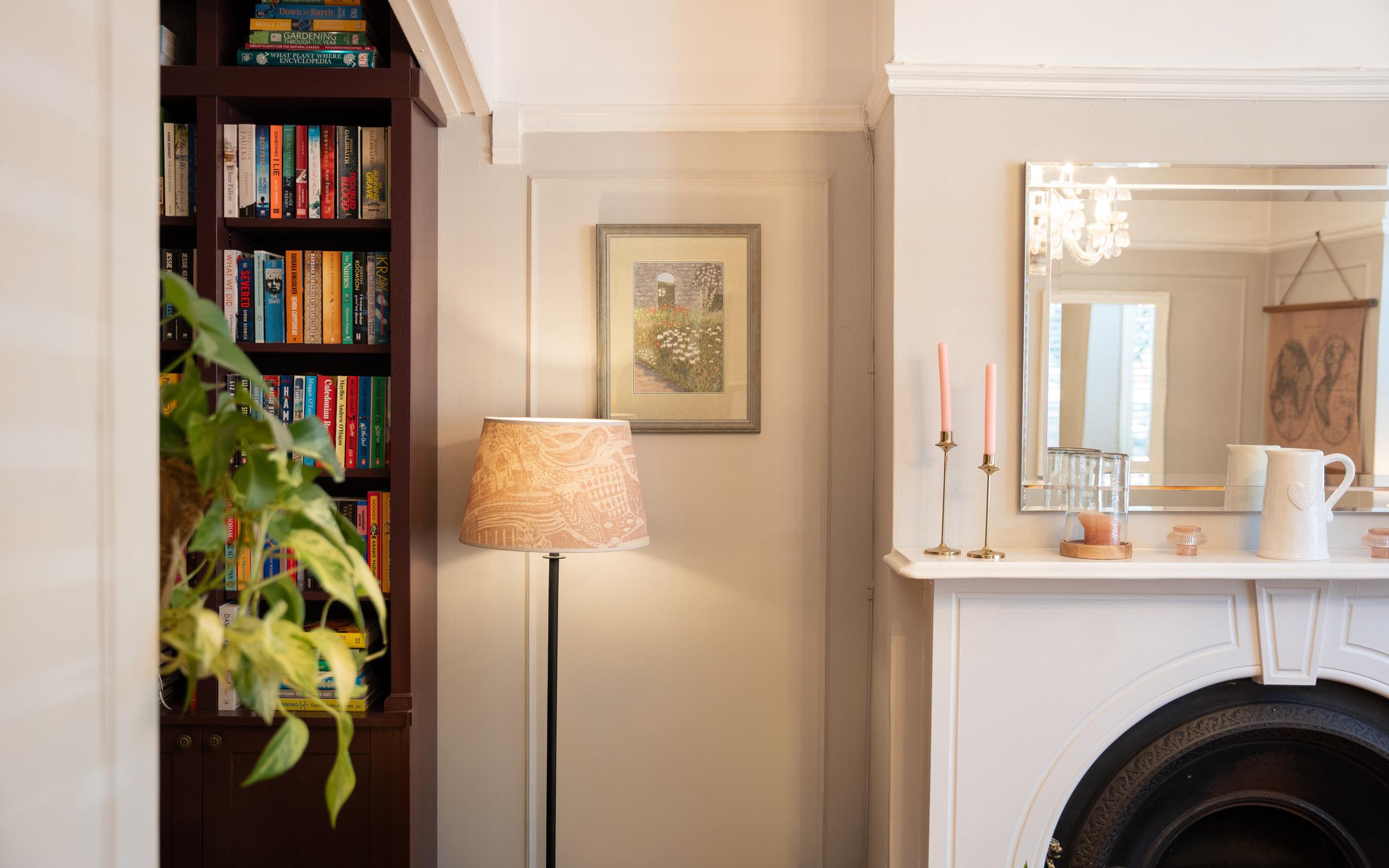 Sitting room with filled bookcase, lit lamp, and white fireplace mantel beneath a mirror