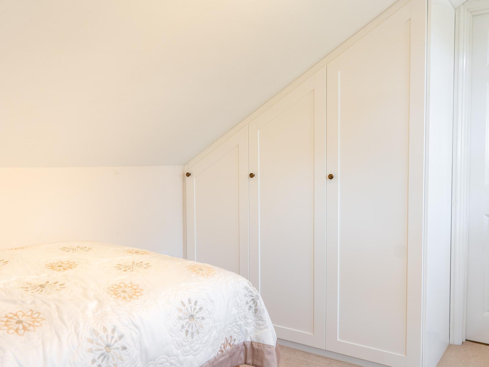 Three white Shaker wardrobe doors stepping down under a sloping bedroom ceiling, with the foot of a bed in the foreground