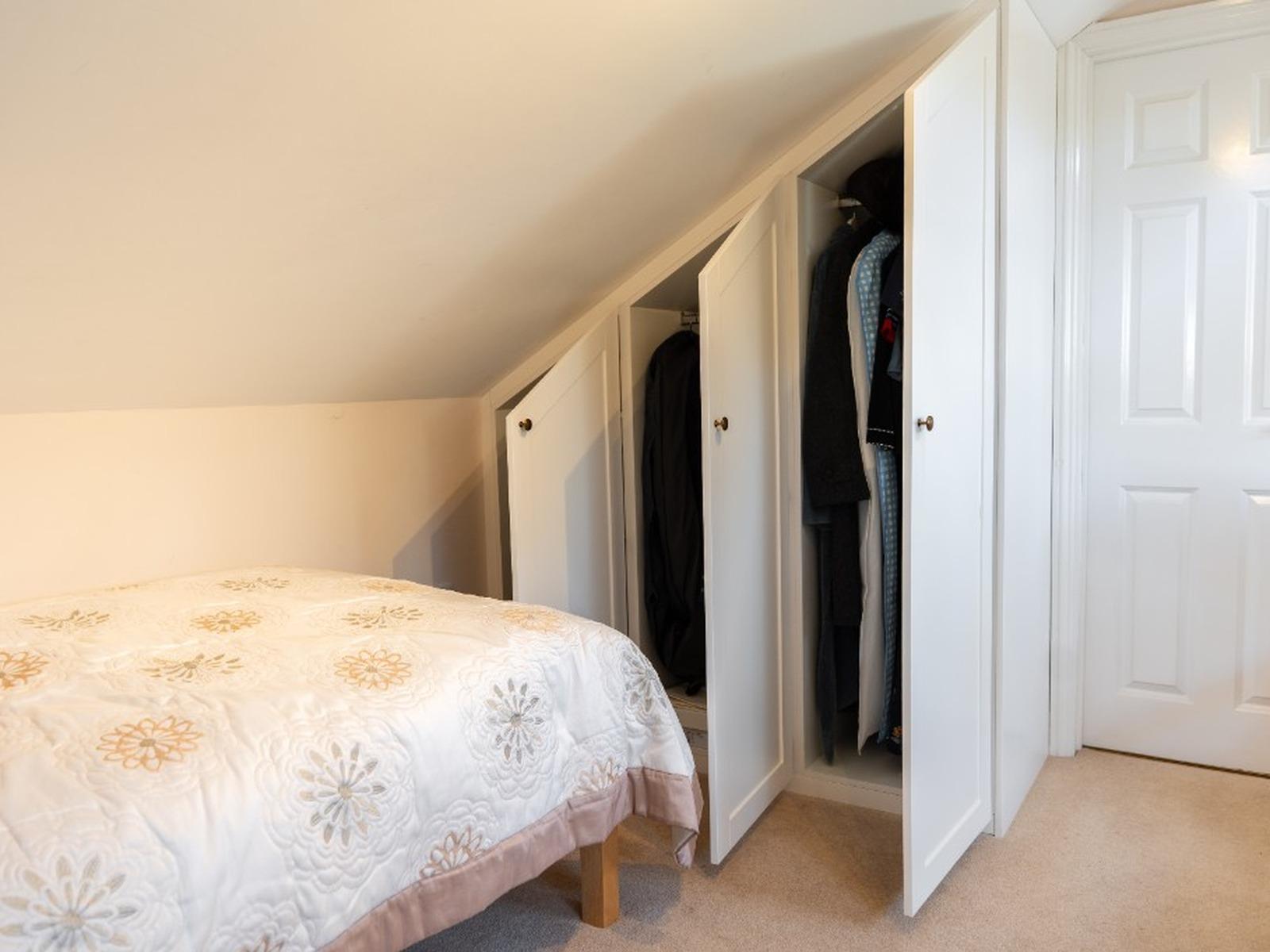 Open white Shaker wardrobe doors under a sloping ceiling, showing hanging rails and clothes