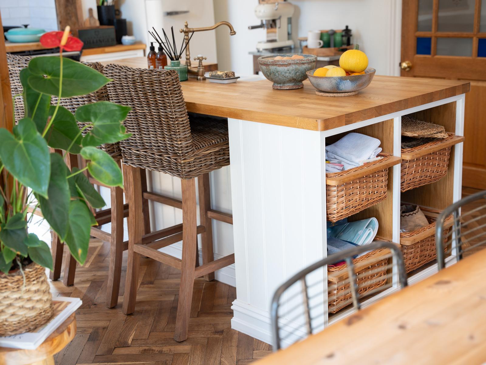 Kitchen island with a wooden worktop, rattan stools, and fitted open cubbies holding woven baskets